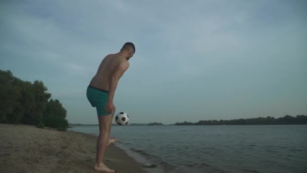 川のほとりにサッカーのハンサムな男 ハンサムな男とともに裸の胴が詰め込まれていますサッカーボールスタンドオンザビーチ — ストック動画