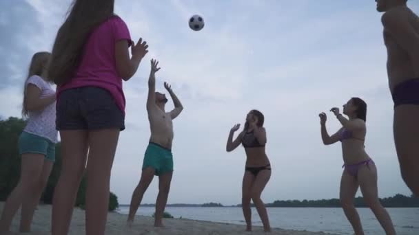 ビーチでボールを持って遊んでいる若い人たち 若い人たちの美しい会社がビーチに立っています 男と女がボールを投げ合う — ストック動画