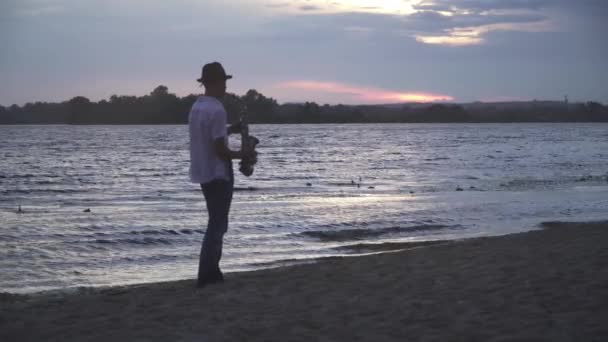 英俊的小伙子正在河边弹奏萨克斯风 穿着白衬衫戴着帽子在海滩上的家伙 背后是美丽的河流 — 图库视频影像