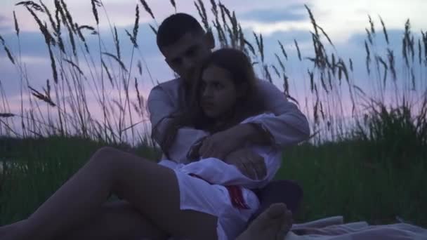 若い男はビーチで美しい女の子を包含します 民族衣装を着た男が美しい少女を抱きしめる 若いカップルは川の近くのビーチに座っている 背景に高い草 — ストック動画
