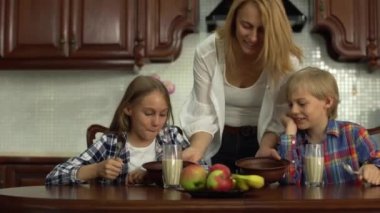 Mutlu anne çocukları sabah okuldan önce besliyor. Gülümseyen kardeşler mısır gevreği yiyor ve mutfaktaki masada süt içiyor. Çocukları öpen kadın. Mutlu dost aile kahvaltılarının tadını çıkarıyor.