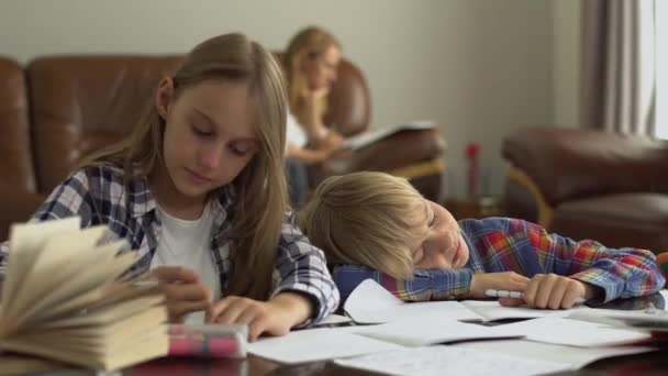 母親が背景に座っている間 かわいい男の子と女の子は前景に家で勉強します 兄は眠りに落ち 妹は彼を目覚めさせた 子供たちは一緒に宿題をする — ストック動画