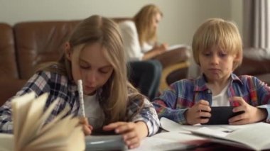 Küçük şirin bir çocuk ve kız evde ön planda çalışıyor. Cep telefonuyla internetten cevapları kontrol ediyor. Çocuklar ödevlerini yapıyor.