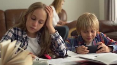 Sevimli kız ve erkek çocukları ön planda çalışıyor. Anneleri arka planda otururken internetten cevapları kontrol ediyorlar. Çocuklar ödevlerini birlikte yapıyorlar.