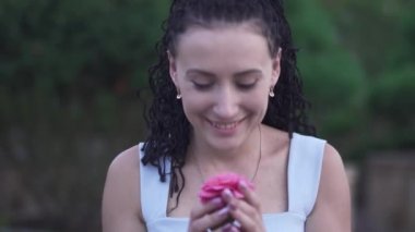 Yetişkin, güzel, beyaz elbiseli esmer kadın bahçedeki çiçeğin kokusunu alıyor. Kadın kameraya çiçek uzatarak elini uzatıyor. Yeşil Bahçe 'de zaman doluyor. Kadın gülümsüyor ve mutlu.