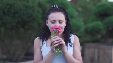 Yetişkin, güzel, beyaz elbiseli esmer kadın bahçedeki çiçeğin kokusunu alıyor. Kadın kameraya çiçek uzatarak elini uzatıyor. Yeşil Bahçe 'de zaman doluyor. Kadın gülümsüyor ve mutlu.