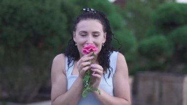 Yetişkin, güzel, beyaz elbiseli esmer kadın bahçedeki çiçeğin kokusunu alıyor. Kadın kameraya çiçek uzatarak elini uzatıyor. Yeşil Bahçe 'de zaman doluyor. Kadın gülümsüyor ve mutlu.