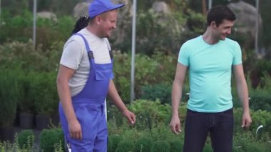 Mavi üniformalı uzun bahçıvan bahçedeki müşterilere beyaz çiçek gösteriyor. İnsanlar mutlu ve gülümsüyor. Erkek ve kadın çiçek kokusu alır.