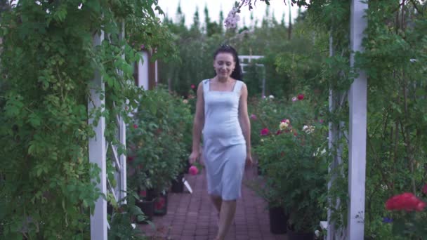 身穿白衣 头戴花环的美丽的成年黑发女士在花园里散步 女人看着天空 在绿色花园等待户外时间 女人又笑又开心 — 图库视频影像