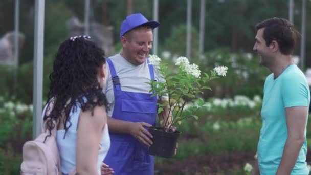 青い制服の背の高い庭師は庭の顧客に白い花を示しています 人々は幸せと笑顔です 男と女の匂いの花 — ストック動画