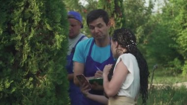 İşçiler ağacın durumunu inceliyor. Bir kadın ve iki erkek bitkiler hakkındaki bilgileri not alıyor. Bahçıvanlar işlerini dikkatli yaparlar.