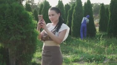 Bahçıvanlar ağaçlarla ilgilenir. Kadın dalların fotoğrafını çeker ve erkek şekil vermek için yaprakları keser. İnsanlar işlerini dikkatli yaparlar. Kamera sola hareket ediyor.