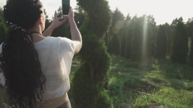 Güzel kız cep telefonuyla bir çalılığın fotoğrafını çekiyor. Bahçe merkezindeki genç kız. Siyah saçlı bir kız parkta fotoğraf çekiyor..