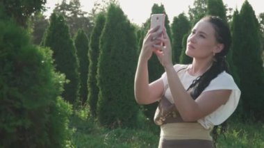 Güzel kız cep telefonuyla bir çalılığın fotoğrafını çekiyor. Bahçe merkezindeki genç kız. Siyah saçlı bir kız parkta fotoğraf çekiyor..