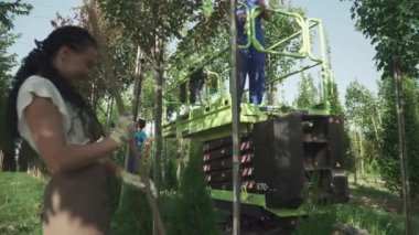 İşçiler bahçede meyve toplarlar. Mevsimsel hasat işlemi devam ediyor. İnsanlar makineli asansörle hasat topluyor. Ağaçlar bahçede düzenli sırada yetişir. Aşağıdan ateş ediliyor.