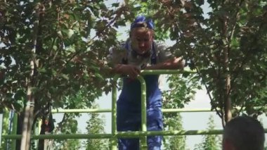 Bahçıvanlar bahçede el sıkışıyor. Bir adam asansörde duruyor, diğeri yerde. Mevsimsel hasat işlemi devam ediyor. İnsanlar makineli asansörle hasat topluyor.