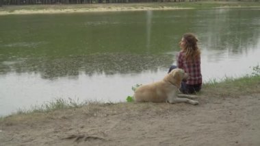 Kıvırcık kadın ve labrador köpeği nehir suyunun yanında oturuyorlar. 