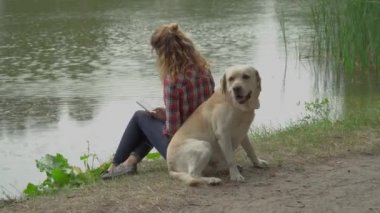 Kafkasyalı kıvırcık saçlı kadın cep telefonu kullanıyor, Labrador köpeği parkta gölde su içiyor, video çekiyor. 