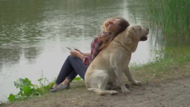 Kıvırcık kadın ve labrador göl kenarında sırt sırta oturuyorlar. Kadın hayvanına yaslanıyor ve akıllı telefon ve video kullanıyor. 