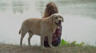 Kıvırcık kadın ve Labrador köpeğinin göl kenarında oturduğu video. 