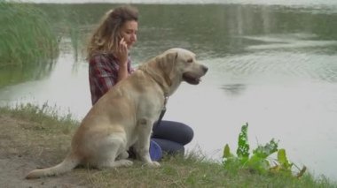 Kıvırcık saçlı kadın ve Labrador köpeği göl kenarında oturuyor. Bayan cep telefonu ve video ile konuşuyor. 