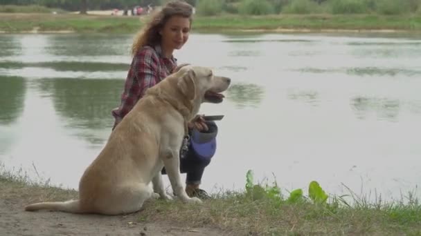 冷たい髪の女性とラブラドール犬は湖の水の近くに休んでいます 女の子が立ち上がって犬を地面に座らせる ビデオ — ストック動画