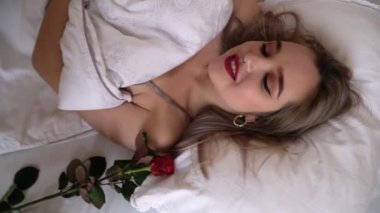 Parlak makyajlı gülümseyen genç bayanın yatakta uzanmış videosu. Kadın kameraya bakarak göz kırpıyor. Kırmızı gül yakınlarda yatıyor. 