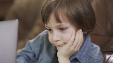 Deri koltukta oturan kot ceketli çocuk dizüstü bilgisayarda mutlu bir şekilde gülümsüyor. Modern çocuğun boş vakitleri. Aygıtlara bağımlılık