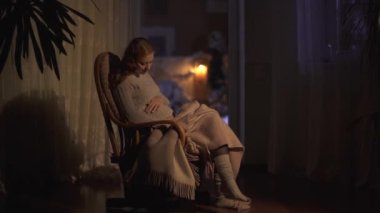 Uzun saçlı güzel hamile kadın karanlık odada sallanan sandalyede oturuyor. Bayan karnını okşa. Kadının bacakları battaniye ve video ile kaplı. 