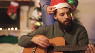 Gitar çalan ve şarkı söyleyen adam, boynunda çelenk olan kadın Noel Baba şapkası takan adam, video. 