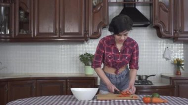 Ekose gömlek giyen, şort giyen, modern mutfağın ahşap tahtasına sebze doğrayan güzel bir genç kız.