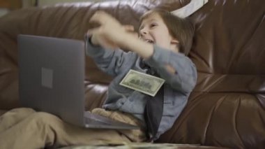 Altı yaşında küçük bir çocuk dizüstü bilgisayarla deri koltukta oturuyor ve havaya para saçıyor. İnternette çalış.