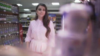 Genç kadın süpermarkette el arabasına canlı kamera koyuyor. Koyu renk saçlı, açık renk elbiseli güzel bir kadın alışveriş yapıyor..