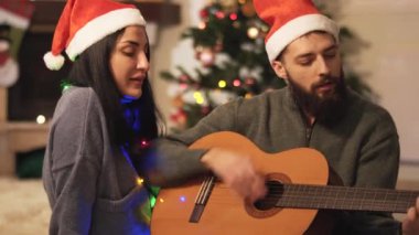 Noel Baba şapkalı mutlu bir çiftin portresi evde şarkı söylüyor. Gitar çalan adam, arka planda Noel ağacı, video 
