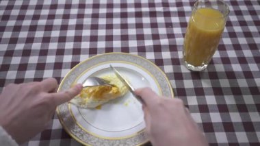Adamın eli tabakta çatal ve bıçakla yumurtayı kesiyor, portakal suyu bardağı kareli masa örtüsüyle kaplı masanın üzerinde. Erkeğin kahvaltısı. Beslenme kavramı. İlk ateş eden kişi.
