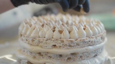 Siyah lateks eldivenli dekoratörün elleri beyaz yuvarlak pastayı kremayla süslüyor.