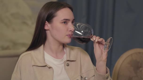 レストランのテーブルに座って大きなワイングラスから赤ワインを飲んでいる長い暗い髪の若い幸せな美しい女性 — ストック動画