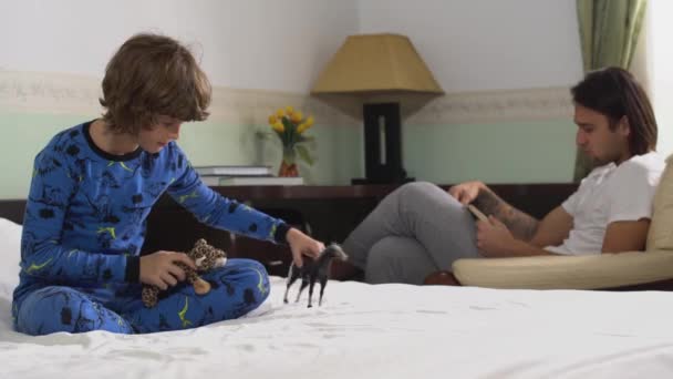 1人の小さなかわいい子供は馬と虎のおもちゃでベッドの上で遊んでいますそして兄で本を読んでアームチェア — ストック動画