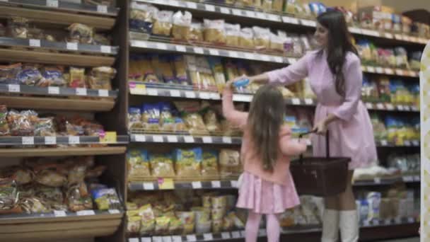 幼い娘はスーパーで買い物をするのに役立ちます スーパーにお母さんがいる女の子 娘はお母さんが店で買い物をするのを手伝う 小さな娘が食料品店で食べ物を買っている美しい暗い髪の母親 — ストック動画