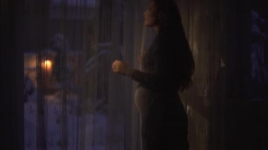 Uzun altın saçlı güzel hamile kadın pencereye gelir ve dışarı bakar. Bayan karnına dokunuyor ve gülümsüyor. Oda karanlık, fener açık havada, video var. 