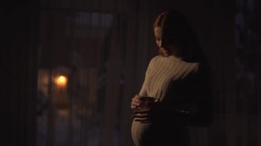 Uzun altın saçlı hamile bir kadın koyu renkli bir odada çay tutuyor. Bayan bir bebek bekliyor. Fener açık havada ışık kaynağı ve video veriyor. 