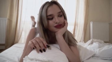 Parlak makyajlı ve burnu delinmiş genç bir kadın büyük pencereli bacakları yukarıda yatakta yatıyor. Kadın kameraya bakıyor ve gülümsüyor. 