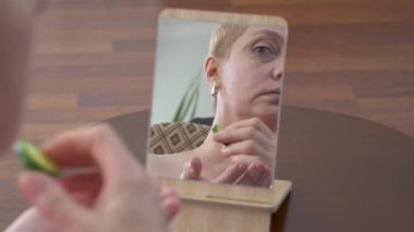 Güzel, yaşlı bir kadının aynaya baktığı ve evinde bir parça salatalıkla pohpohlanan yüzünü gösteren bir video.