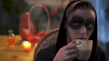 Koyu renk elbiseli genç bir adam sandalyede oturuyor. Siyah içecekler içindeki genç sihirbaz kakao içiyor. Cadılar Bayramı. video