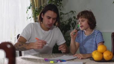 İki erkek kardeş, bir yakışıklı yetişkin ve bir sevimli küçük çocuk masaya oturur ve yakışıklı adam kağıda renkli kalemlerle resim çizer..