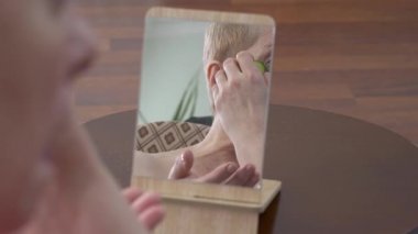 Güzel yaşlı kadının aynaya baktığı ve evinde bir parça salatalıkla yüzünü ovaladığı bir video.