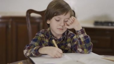 Evde masada oturan küçük yorgun çocuk bir sürü not defteri ve kağıtla birlikte. Çocuk ödev çalışıyor..