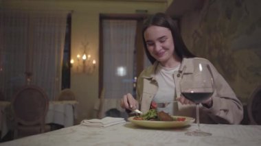 Güzel servis edilen erişteleri roka ile yiyen genç bir kadın kırmızı şarap içip birine gülümsüyor.