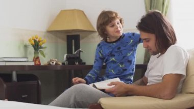 İki kardeş koltukta oturuyor, büyük kardeş küçük kardeşe bir kitap okuyor ve küçük çocuk uzun saçlara dokunuyor ve ağabeyi de saçlarına dokunuyor. Kardeşler birbirleriyle oynarlar..