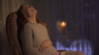 Sallanan sandalyede oturan hamile bir kadın bebeğin hareketini midesinde hisseder ve mutlu bir şekilde güler. 